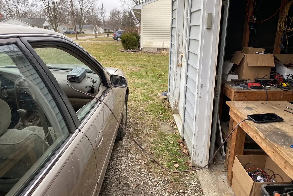 A parked sedan in a gravel driveway with a small inverter on the dashboard and an extension cord running through the cracked window to a tablet on a workbench inside an open shed