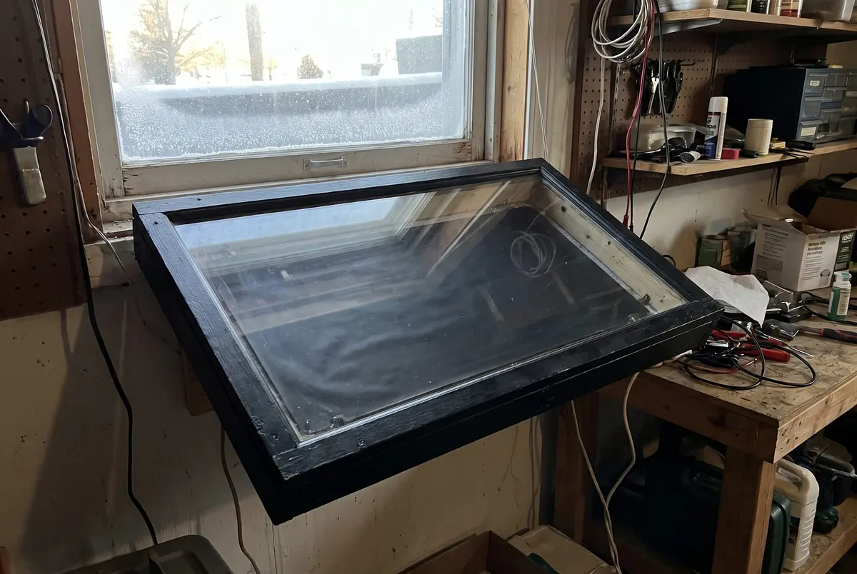 A black wooden box with a glass cover tilted against the bottom of a basement window, with a snowy yard outside and a workbench beside it