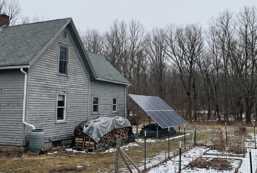 A gray clapboard farmhouse in winter with a rain barrel at the downspout, a covered woodpile and a ground-mounted solar array beside a fenced garden