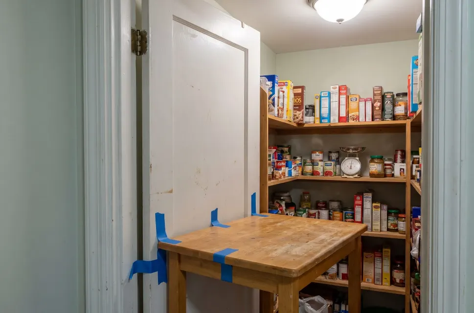 A walk-in pantry with its door open, shelves of canned and boxed food, a kitchen scale on a shelf, and a butcher-block table in front